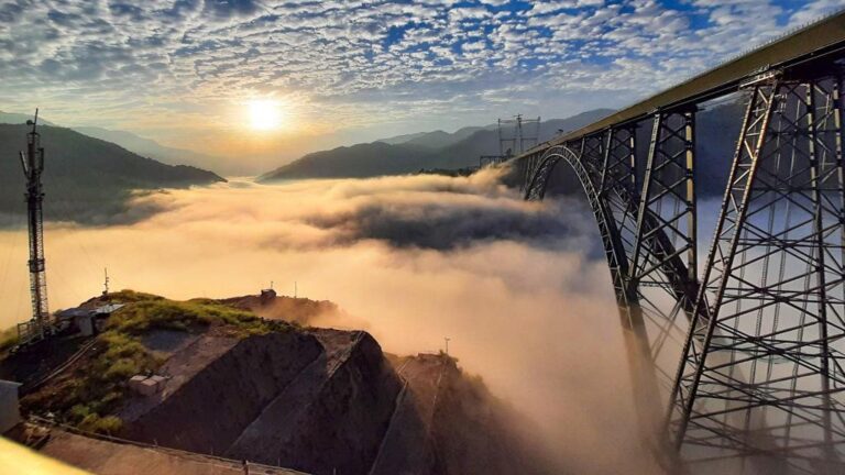Aerial view of the Chenab Rail Bridge, the world’s highest railway arch bridge, emerging through clouds in the Himalayas.